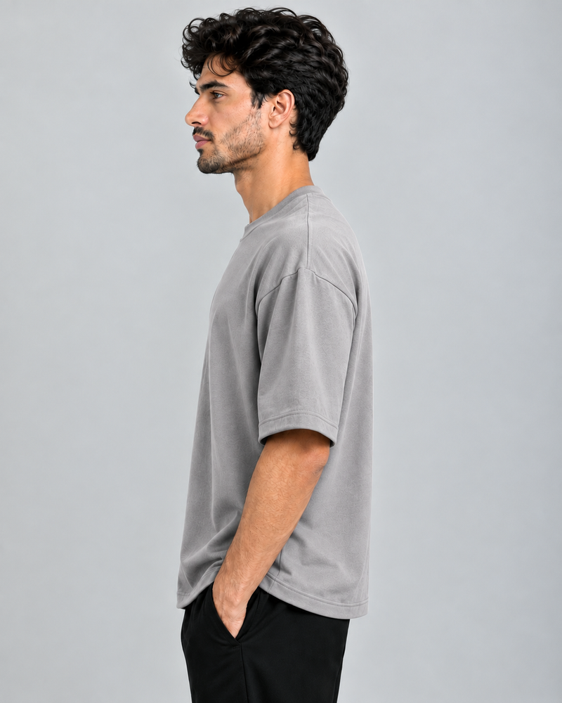 A man in profile wearing an oversized light gray crewneck t-shirt and black pants, standing against a solid light gray studio background.
