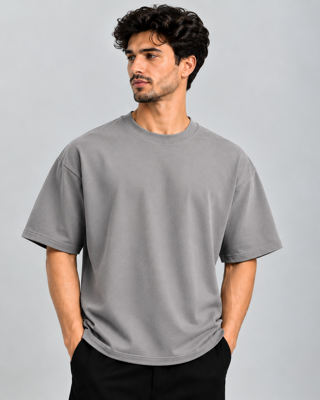 A man wearing a plain gray short-sleeve crewneck t-shirt and black pants, standing against a solid gray studio background.